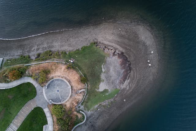 A nadir view of a rounded beach that juts out into the water, with park grass above the shore and paved walking paths that resemble a stick figure with a large round head.