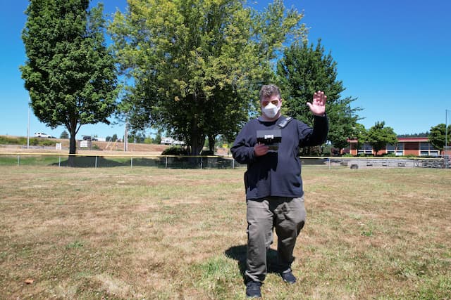 a friendly PIGS staff member waving hello while holding a drone remote control in a baseball field in front of trees.